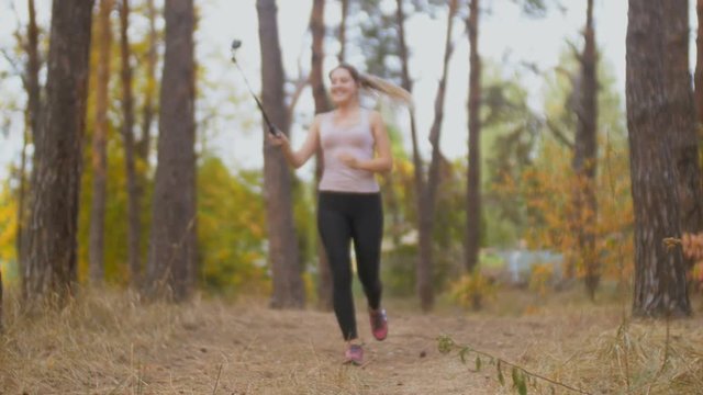 Beautiful smiling woman making video with selfie stick of her running in forest