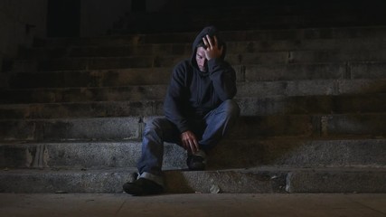4K video lateral panning 24 fps of young desperate wasted man in hood suffering stress and drepression sitting miserable on urban street staircase at night sad and worried in addiction concept