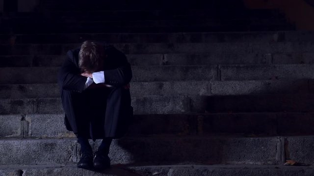 4K video lateral panning 24 fps of young desperate businessman in suit and tie suffering stress and drepression sitting on urban street staircase at night sad and worried in crisis recession concept