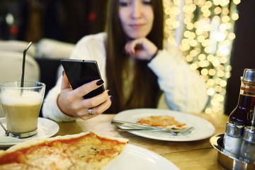 Woman typing write message on smart phone in a modern cafe. Cropped image of young pretty girl sitting at a table with pizza using mobile phone. Tonned. Selective focus.