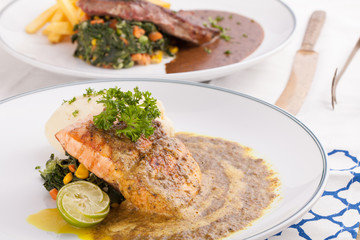 A nice dish of salmon and beef steak served with mashed potato,lemon, saute spinach and sauce on background with wooden utensils.