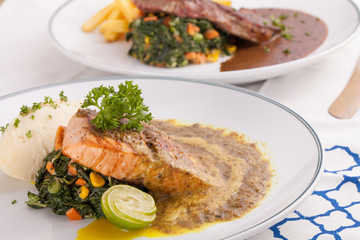 A nice dish of salmon and beef steak served with mashed potato,lemon, saute spinach and sauce on background with wooden utensils.