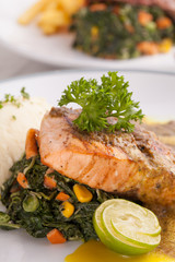 A nice dish of salmon and beef steak served with mashed potato,lemon, saute spinach and sauce on background with wooden utensils.