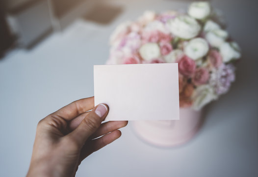Close-up of female hand holding blank craft business card with empty space for your design or logo, hat box of flowers in the background, mock-up of paper business card