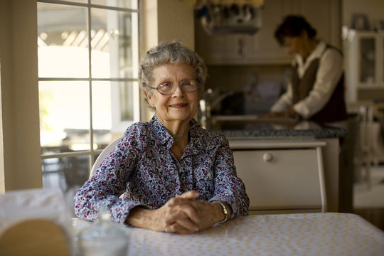 Portrait of a cheerful elderly woman sitting at her kitchen table while her daughter helps out with the dishes.