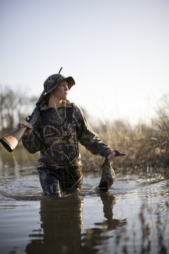 Young woman walking through a fiver carrying a shotgun and dead duck.