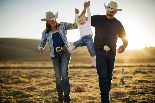 Farmer and his wife swinging their daughter into the air between them on their ranch at sunset.