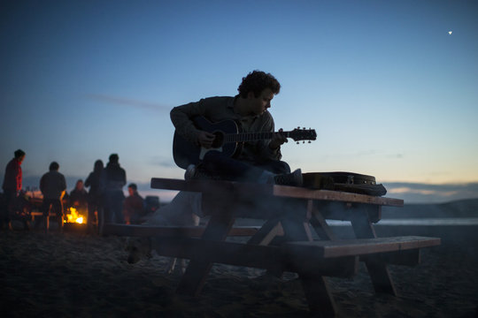 Guitar player sitting on a picnic table at the beach at sunset.