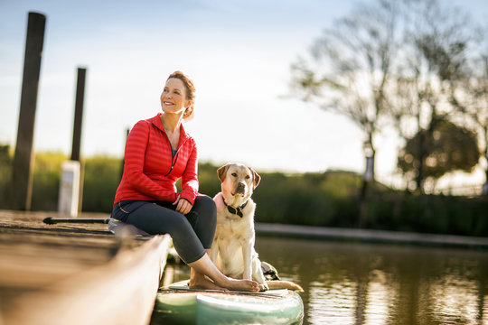 Mid adult woman sitting on wharf with pet golden labrador.
