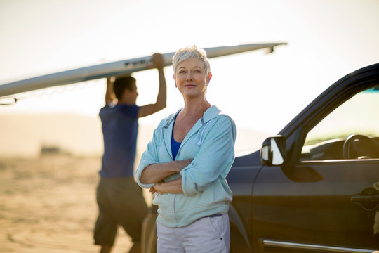 Portrait Of Happy Woman Standing By Her Car While Man Lifting Surfboard