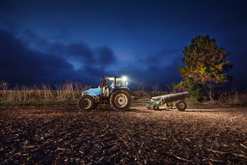Naklejka premium Tractor preparing land with seedbed cultivator at night