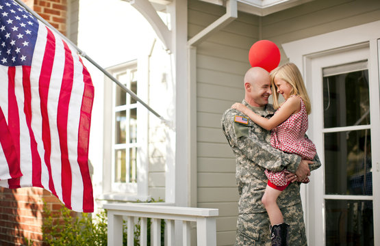 Male soldier hugging his young daughter on the porch of their home.