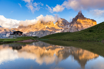 The Pale di San Martino peaks (Italian Dolomites) reflected in the water at sunset, with an alpine chalet on background. - 134387419