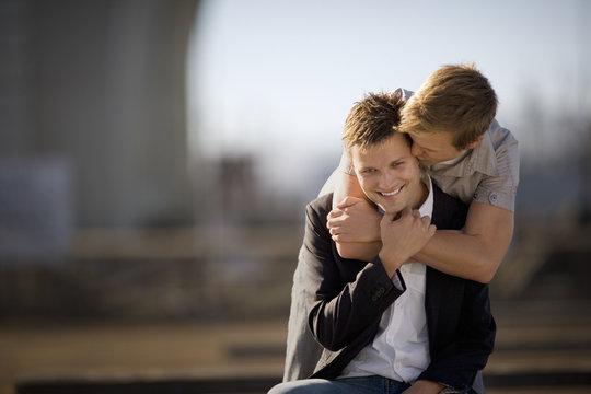 Young man giving his boyfriend a kiss on the ear while standing with his arms around him.