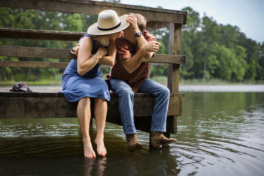 Mature man romantically leans in to kiss his wife as they sit together on a wooden pier over a lake.