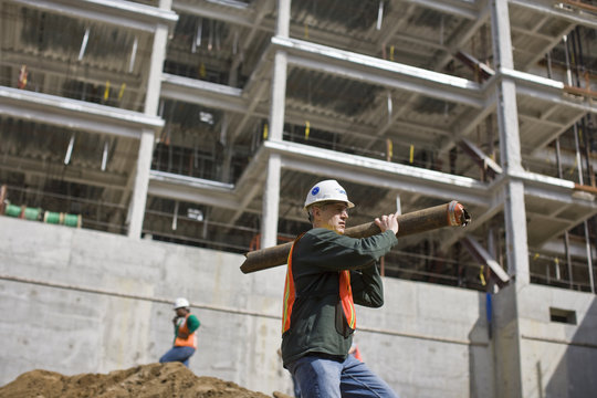 Construction worker wearing a hard hat and carrying a large pipe on an outdoor construction site.