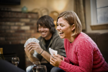 Young woman laughing while holding a hand of playing cards inside a sitting room with friends.