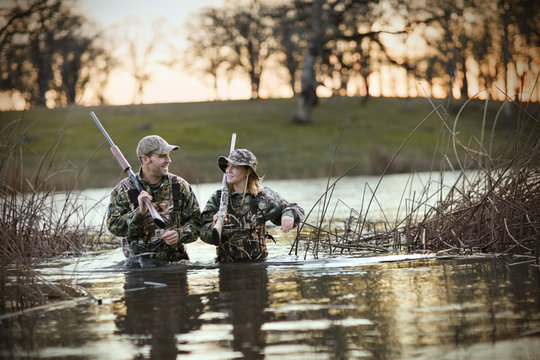 Male and female hunter wading through a lake carrying rifles