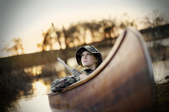 Young adult woman sitting in a canoe with a gun by a lake.