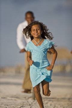 Portrait of a smiling young girl in a blue dress running along a sandy beach followed by her father on a bicycle.