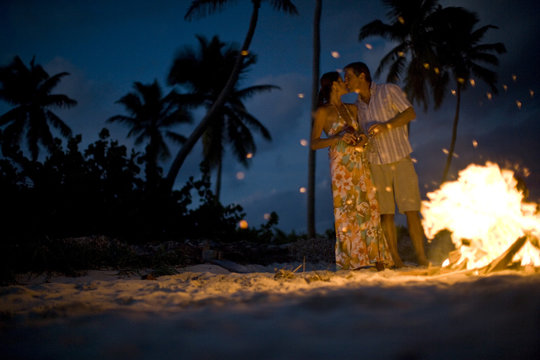 Mid-adult couple standing beside a bonfire on a beach.
