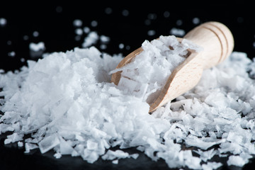 Salt flakes and wooden scoop on black background