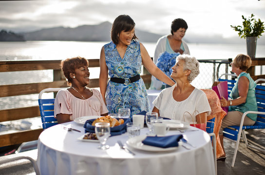 Two mature female friends out to lunch at a seaside restaurant share a laugh with an acquaintance who has come to say hello.