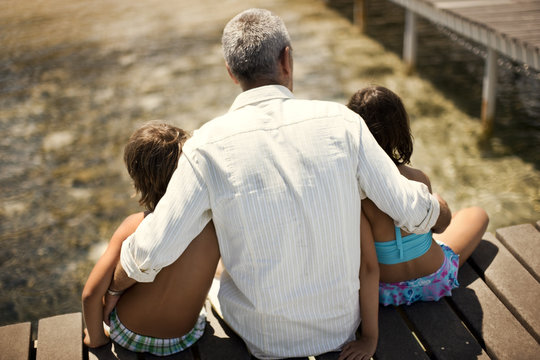 Old grandfather sitting with his two young grandchildren on a wooden jetty at the beach.