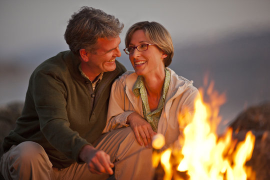 Smiling middle aged couple sitting by a bonfire on a beach.