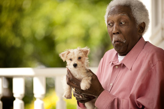 Senior man makes a face as he holds a puppy in his hands as he stands on a porch.