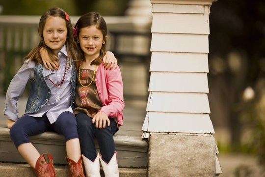 Two young girls with their arms around each other sit together on the top step of porch stairs and smile as they pose for a portrait.
