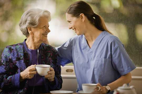 Female nurse puts a reassuring arm around the shoulders of a senior woman as they have tea.