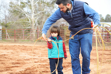 Father training small toddler boy son making loope and throwing lasso to bull in the ranch. Roping simulator. Red clay background, countryside. Family, relationship, free time