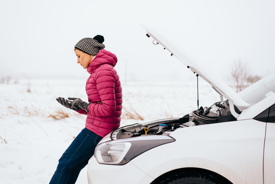 Young Woman Calling For Help Or Assistance After Her Car Breakdown In The Winter. Broken Down Car With Open Hood On A Country Road.