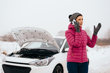 Young woman calling for help or assistance after her car breakdown in the winter. Broken down car with open hood on a country road.