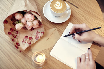 Fototapeta na wymiar Woman hands close-up writing in notebook, using pencil, blank pages for layout. Cappuccino latte coffee with heart on top. Flowers on wooden table in modern cafe. St. Valentines celebration concept