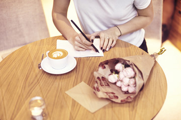 Obraz na płótnie Canvas Close-up hipster businesswoman wearing white t-shirt writing in cafe on startup project. Creative young girl using notebook at wooden table. Drinking coffee break. 