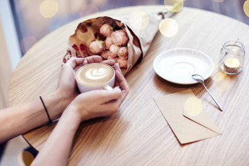 Cup of coffee with heart in woman hands, envelope and flowers on wooden table in cafe. bokeh on background. Focus on left hand. 