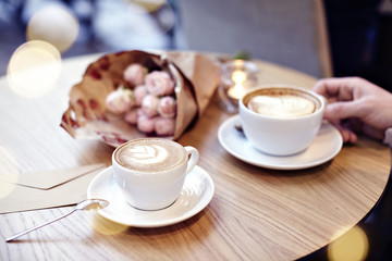 Two Cups of coffee with heart and flowers on wooden table in cafe. Man hand holding a cup. Bokeh on background. Focus on coffee cup.