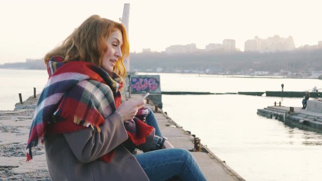 Young woman with red hair curls sitting in a scarf and using phone at the seacoast