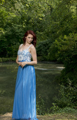 Obraz na płótnie Canvas Portrait of stunning red haired woman wearing a blue dress standing in shade of wooded area near pond