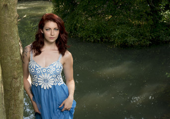 Portrait of stunning red haired woman wearing a blue dress standing in shade of wooded area near pond