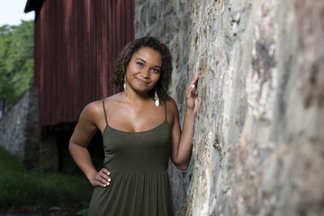 Stunning young biracial woman in green sundress standing next to covered bridge and stone wall foundation - rural