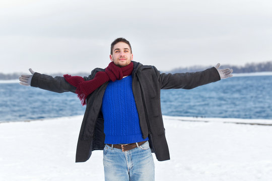 Man opened his arms to the sides. He is dressed in coat, scarf, jeans and gloves. Winter.