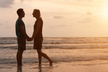 Fototapeta premium Couple in love is staying on beautiful sunset beach, holding their hands and looking on each other. Love and romance in vacation honeymoon.