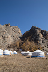 White and blue canvas gers at the base of rugged mountains. The yurts are on stone foundations. Sparse vegetation is in the background. Deep blue sky.