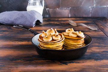 Stack of homemade pancakes with banana, maple syrup and walnuts in black cast iron skillet. Vintage forks, glass jar with milk, wooden rustic table.
