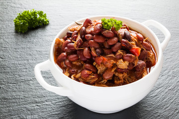 Beans with pork stewed in spicy tomato sauce with onion, paprika, beer, bell and pink pepper. White soup bowl and fresh parsley on black stone.