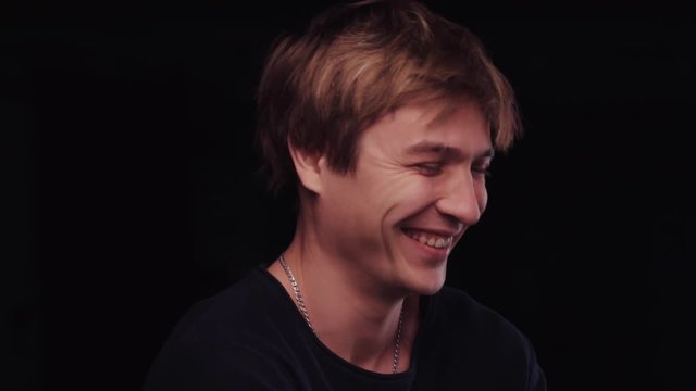 Close up view of handsome young man in a casual black sweater. Attractive guy is laughing, not looking forward to the camera. Happiness, being funny. Male portrait, black background, inside shooting.