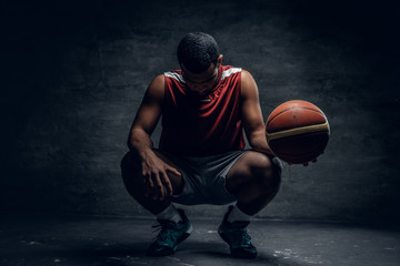 A black basketball player sits on a floor and holds basket ball.